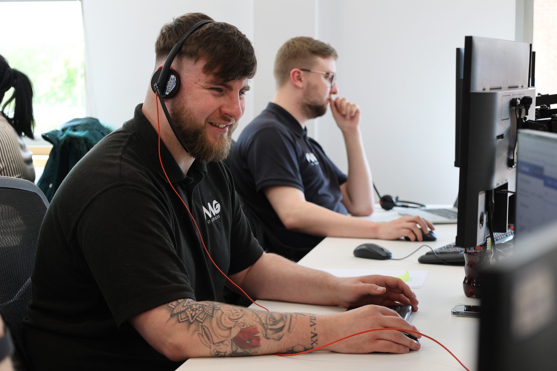 AAG IT Services Technicians Managed IT Support technician working at a desktop workstation in a modern office environment, wearing a headset and using a computer to assist customers with IT issues. Multiple team members are seated at adjacent desks with dual-monitor setups, demonstrating an active IT support desk providing remote troubleshooting, technical assistance, and day-to-day IT management services for businesses. Bright, professional workspace with technology-focused staff delivering responsive managed IT support and customer service.